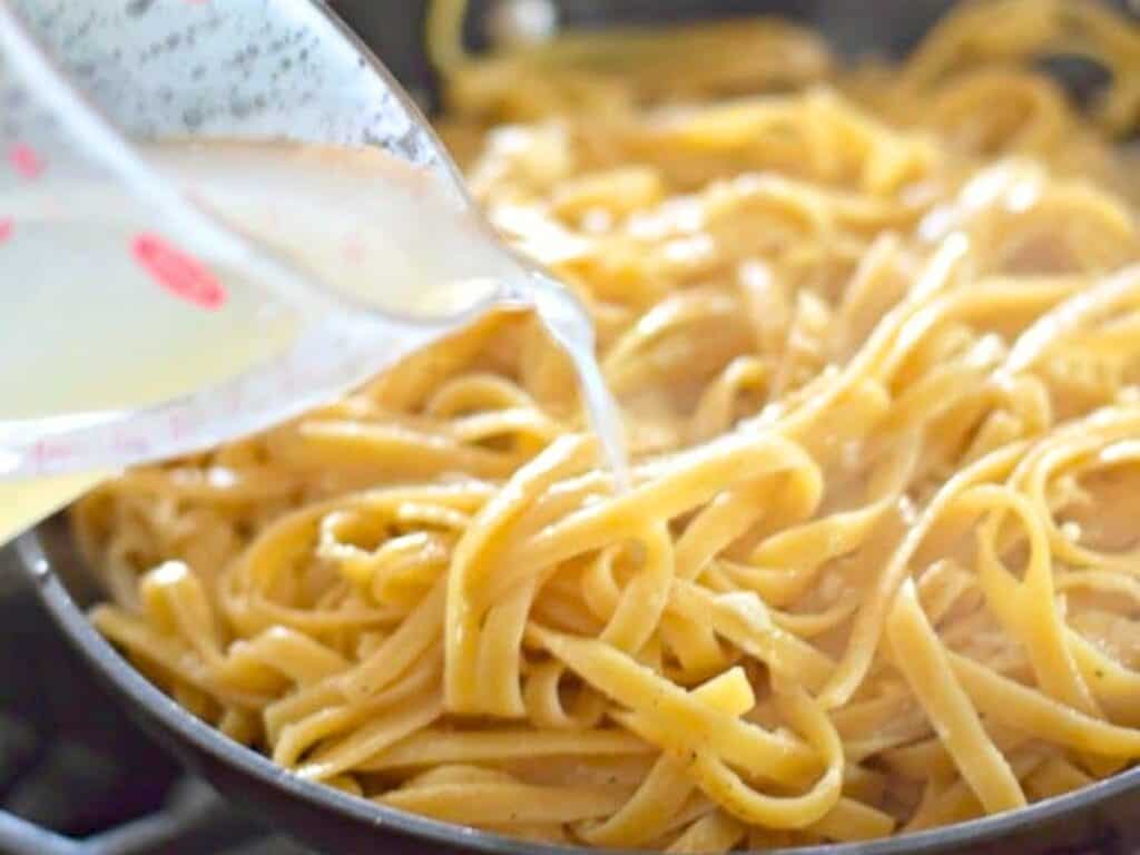 A measuring cup pours liquid onto cooked fettuccine pasta in a pan on a stovetop, creating the perfect base for a creamy cacio e pepe.