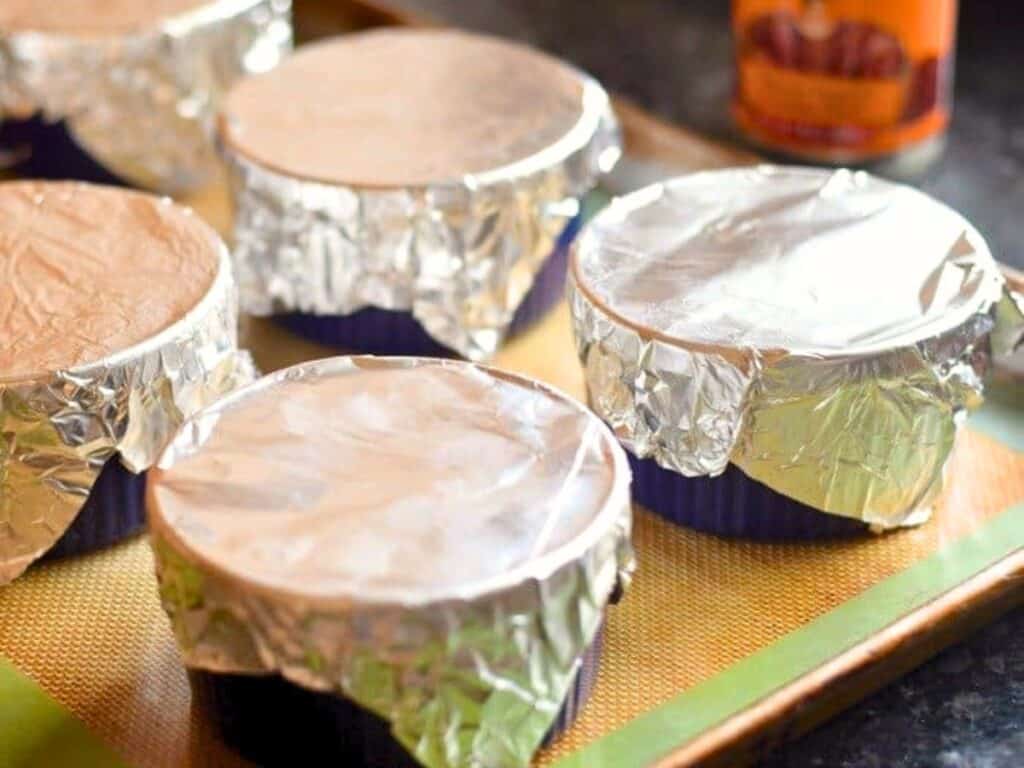 Several round ramekins covered with aluminum foil are arranged on a baking sheet, ready to be baked into savory tamale pot pies.