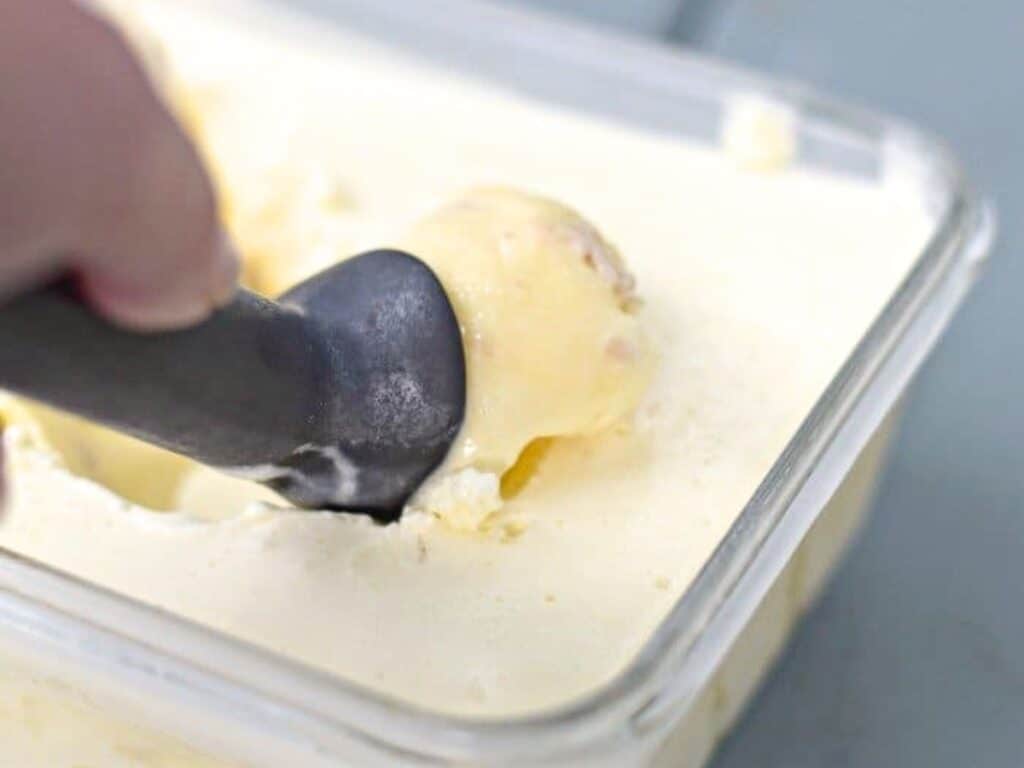 A hand uses an ice cream scoop to serve a portion of homemade frozen custard from a rectangular container.
