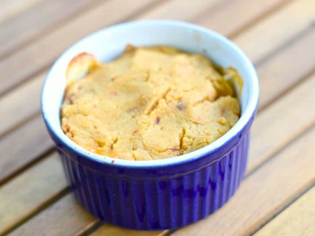 A baked dessert with a golden crust in a round, purple ramekin sits on a wooden slatted table, reminiscent of individual tamale pot pies in both charm and presentation.