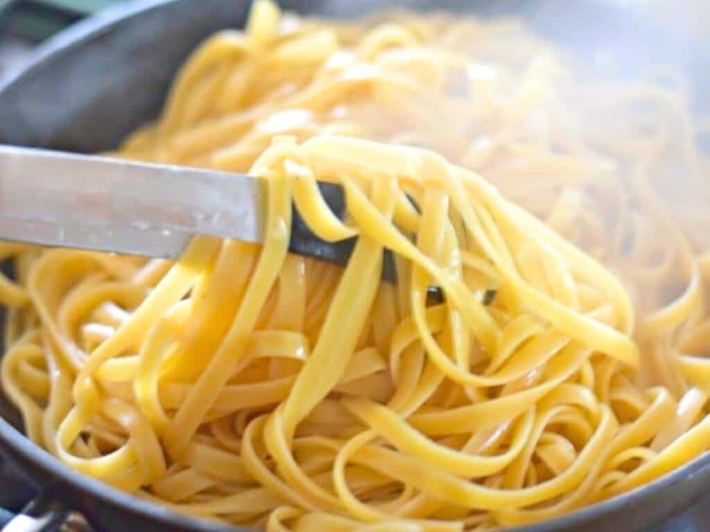 A close-up of cooked fettuccine pasta, perfect for cacio e pepe, being lifted from a pot with metal tongs, with steam rising in the background.