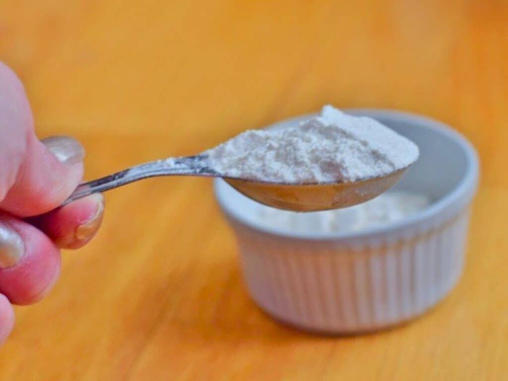 A hand holds a spoonful of white powder above a white ramekin filled with the same powder on a wooden surface, ready for the next step in making a delicious microwave chocolate chip cookie.