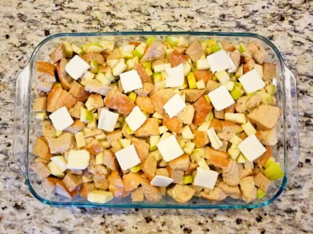 Glass baking dish filled with cubed bread, chopped apples, and evenly spaced pats of butter on a granite countertop—perfect for creating a cozy apple pie bread pudding.