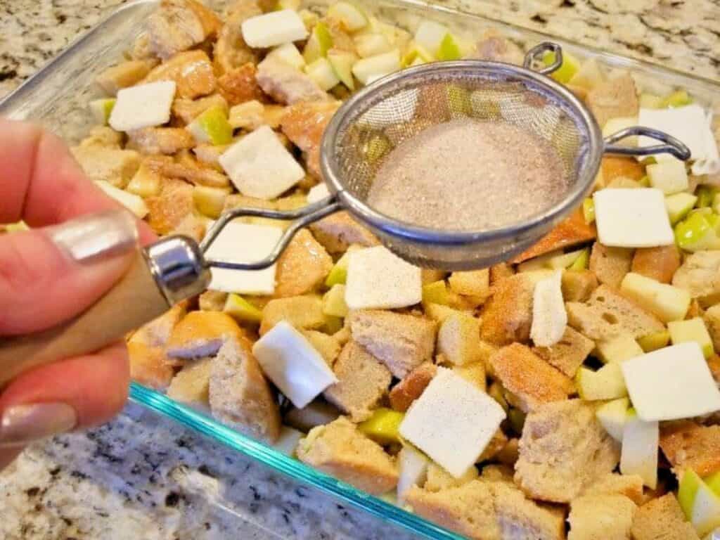 A hand holds a sifter sprinkling cinnamon sugar over a glass dish filled with cubed bread, apples, and butter on a kitchen counter, creating the perfect base for an apple pie bread pudding.