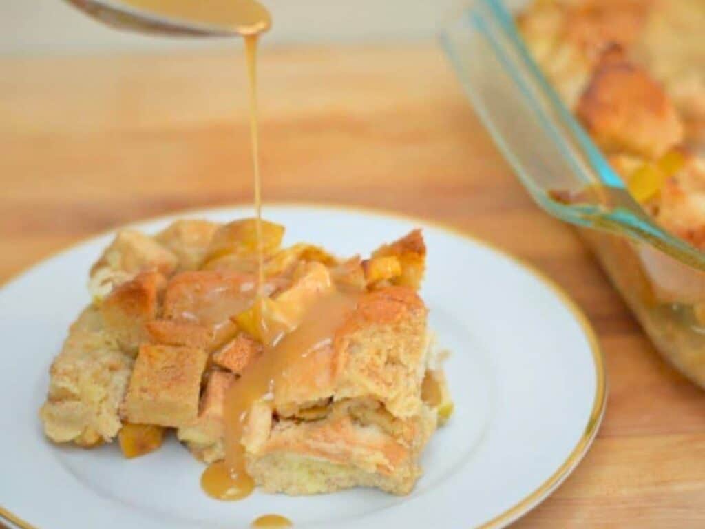 A slice of apple pie bread pudding on a plate is topped with caramel sauce being poured from a spoon, with a baking dish of apple pie bread pudding in the background.
