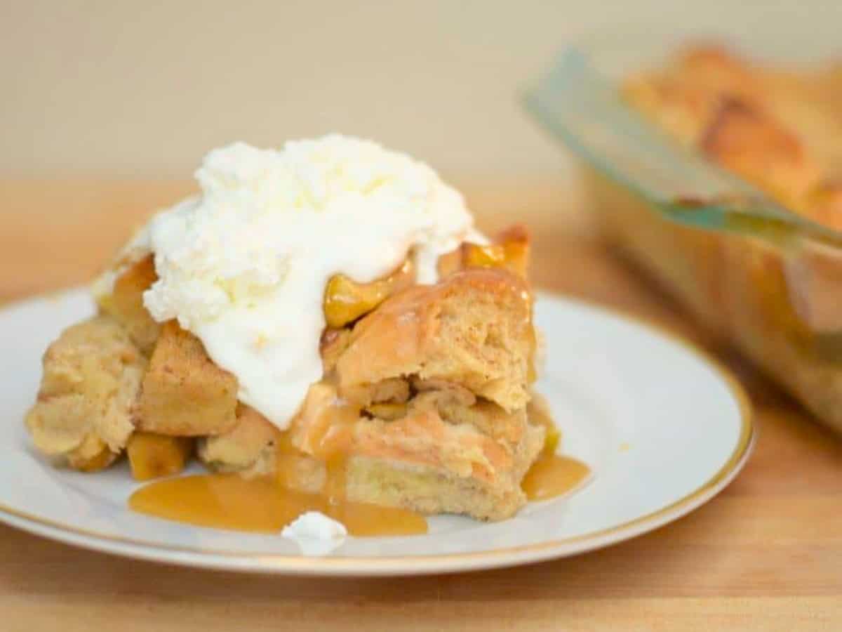 A slice of apple pie bread pudding with caramel sauce and whipped cream on a white plate, with a glass baking dish in the background.