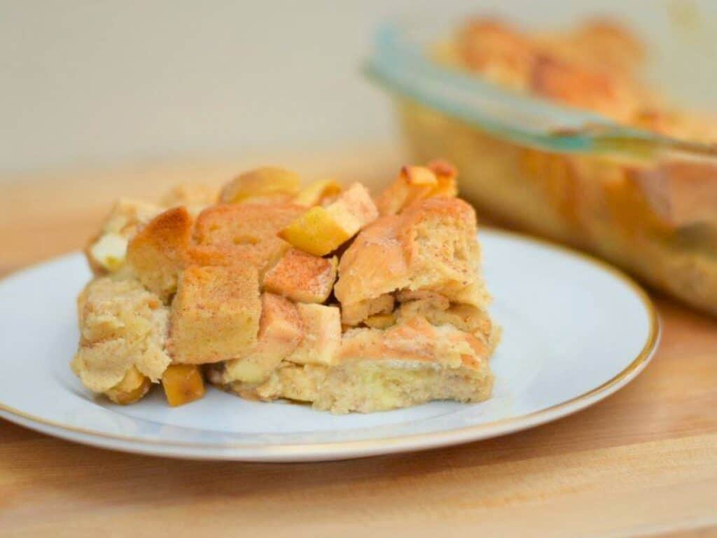 A slice of apple pie bread pudding with visible apple chunks sits on a white plate; a glass baking dish with more is in the background.