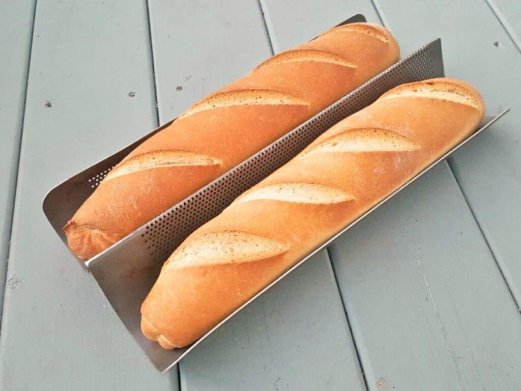 Two golden brown baguettes, crafted from a classic baguette recipe, rest in a perforated metal baking tray on a light gray wooden surface.