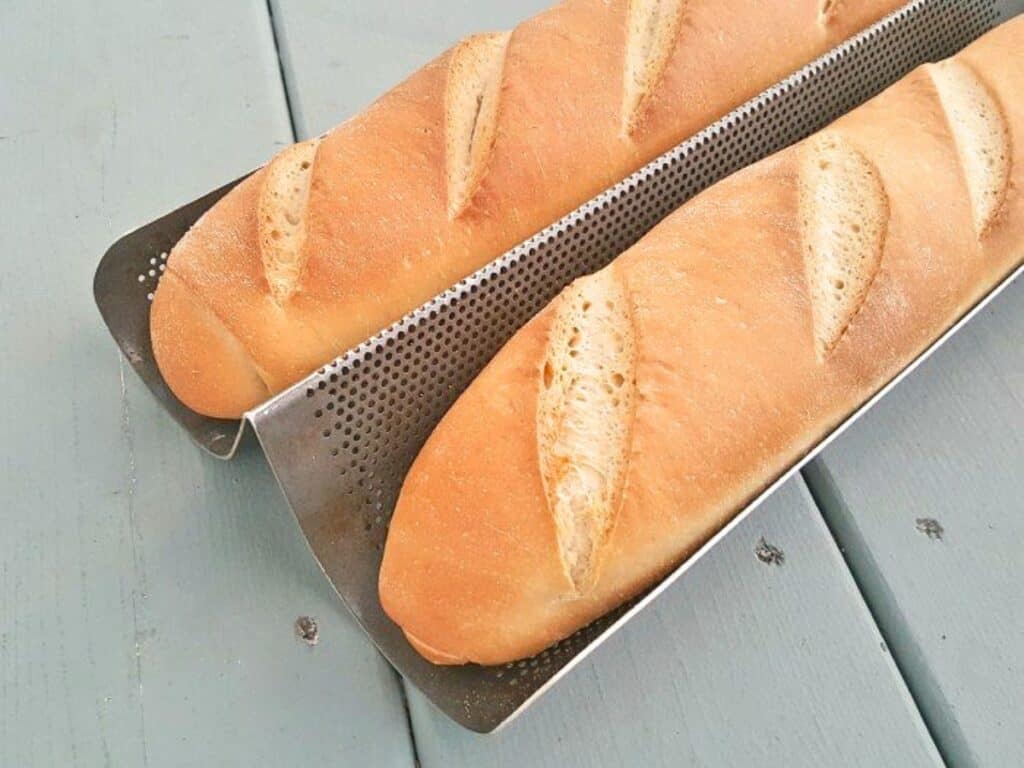 Two freshly baked baguettes with slits on top, made from a classic baguette recipe, are resting on a perforated metal baking tray placed on a light blue wooden surface.