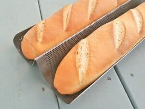 Two freshly baked baguettes with slits on top, made from a classic baguette recipe, are resting on a perforated metal baking tray placed on a light blue wooden surface.