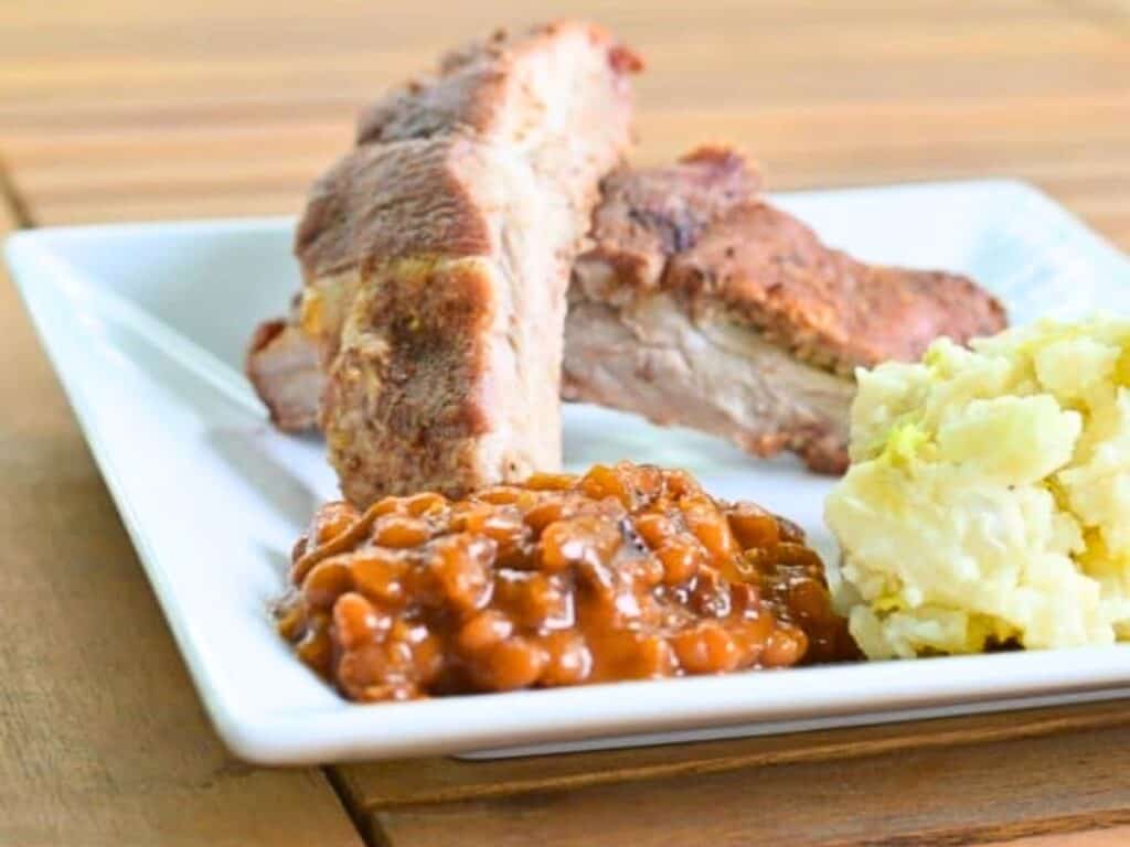 A white plate with Instant Pot baked beans, mashed potatoes, and two pieces of grilled pork ribs sits on a wooden table.