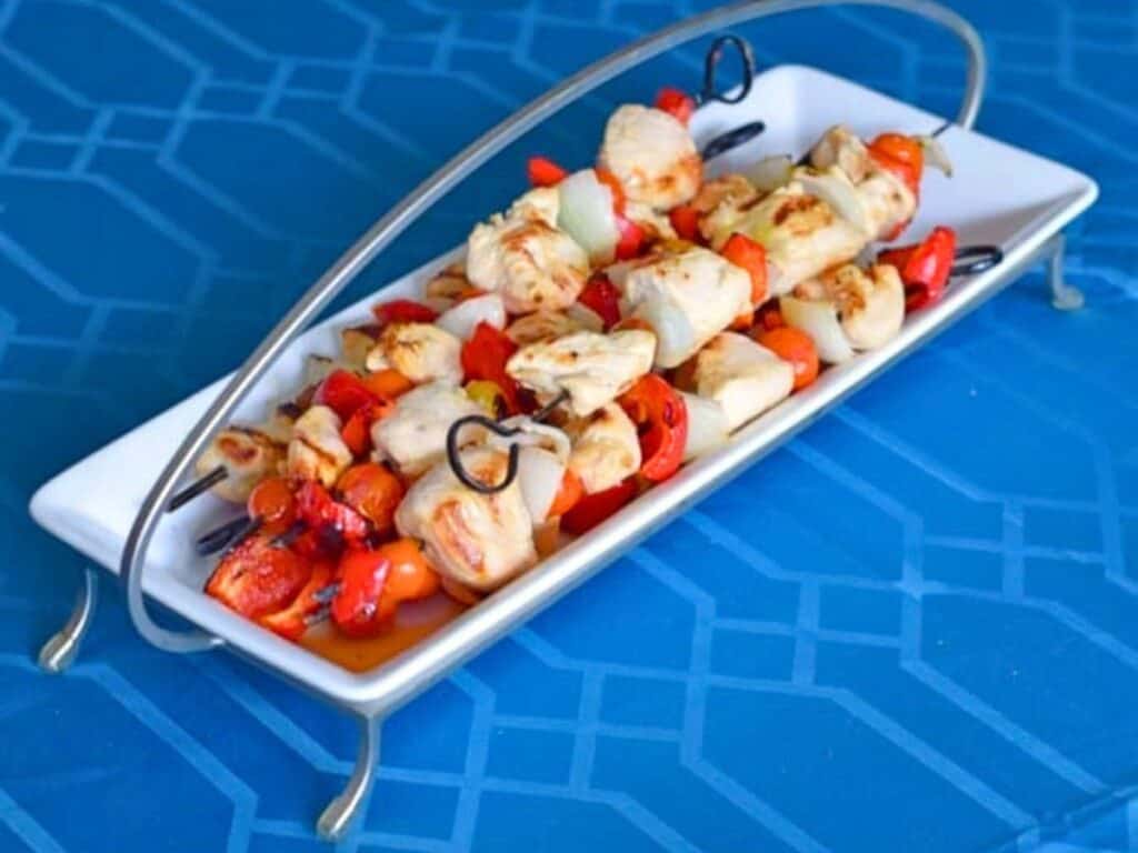 Two skewers with lemon marinated chicken kebabs, onions, and red bell peppers are served on a rectangular white tray with metal handles, placed on a blue patterned surface.