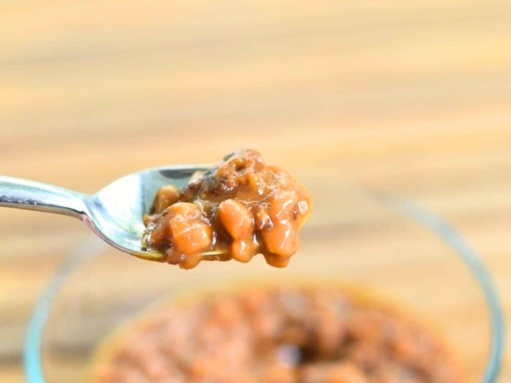 A close-up of a spoonful of Instant Pot baked beans in rich sauce, held above a glass bowl filled with more baked beans.