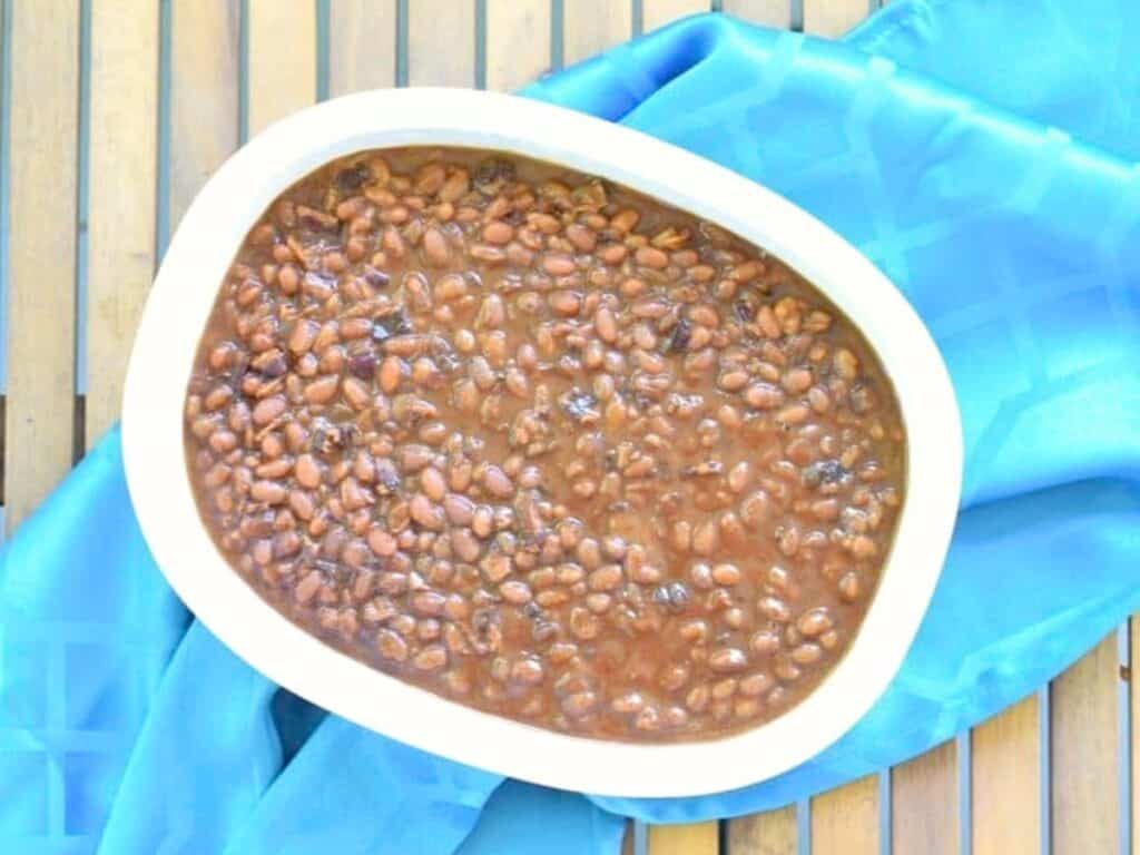 Oval white dish filled with Instant Pot baked beans in sauce, placed on a blue cloth on a wooden slatted surface.