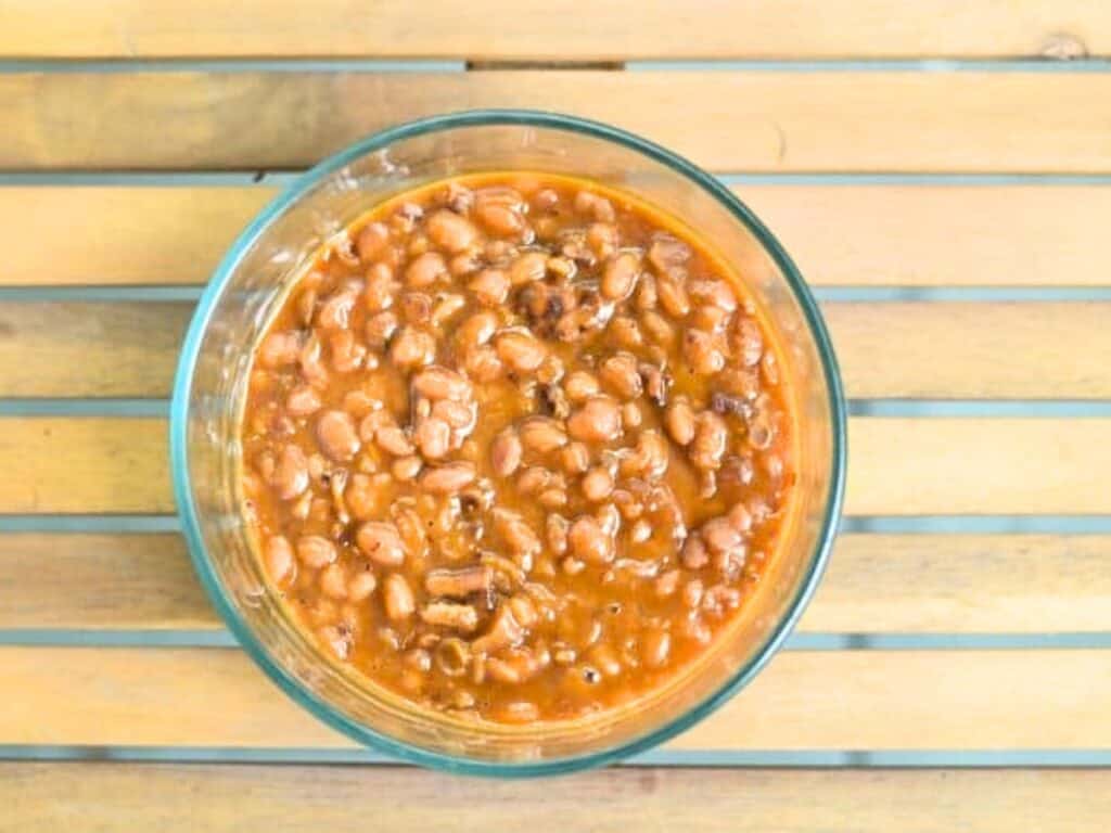A glass bowl filled with Instant Pot baked beans in sauce sits on a wooden slatted surface.