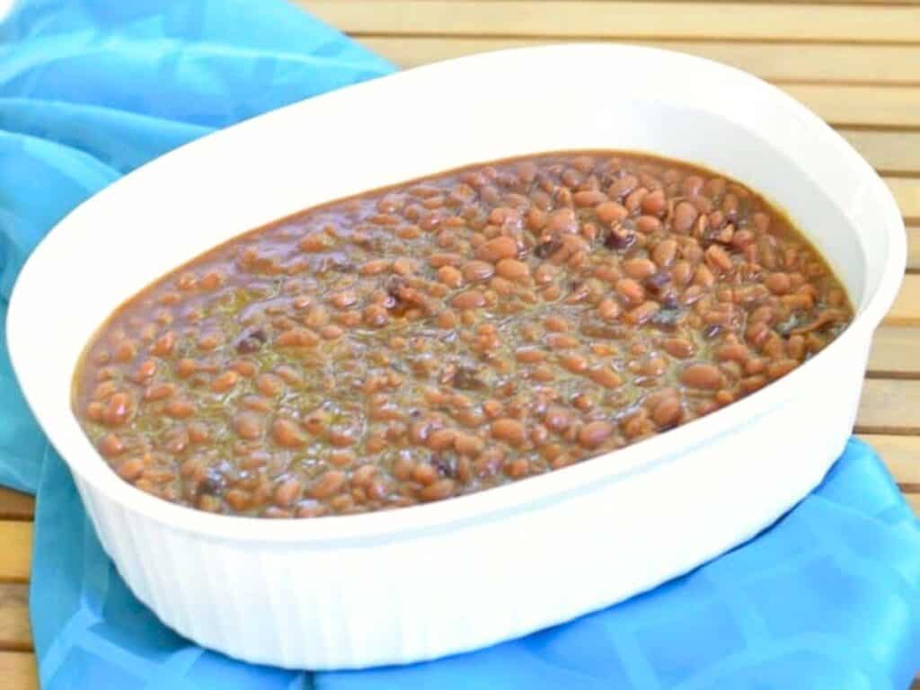 A white casserole dish filled with Instant Pot baked beans sits on a blue cloth on a wooden surface.