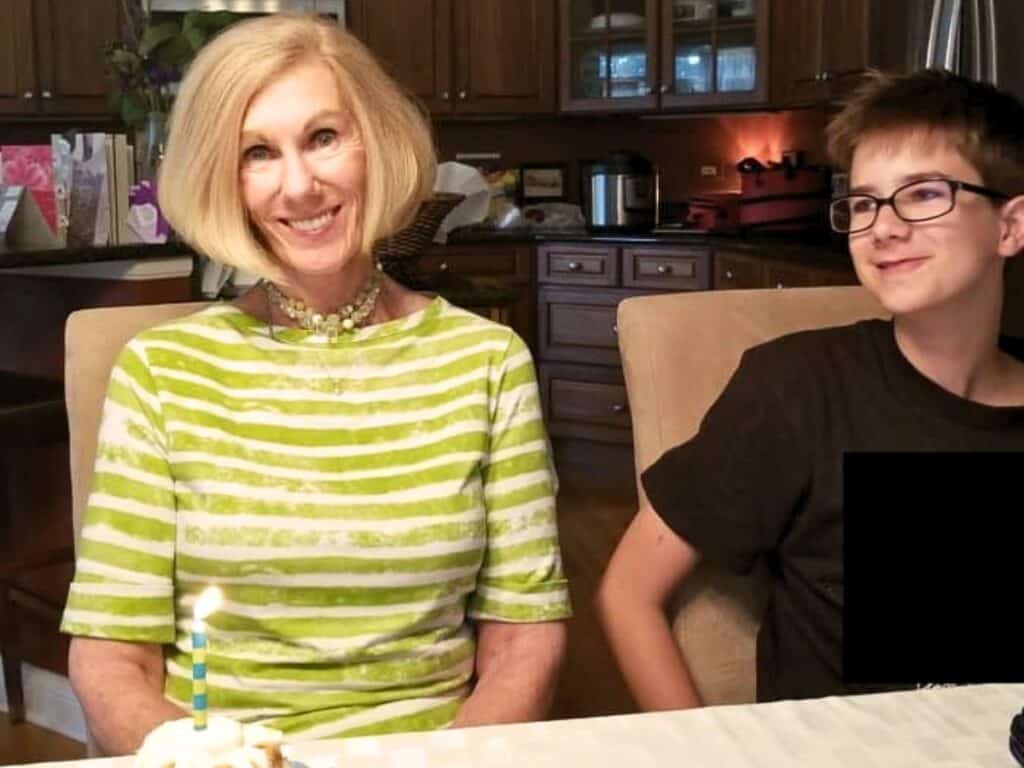 An older woman and a boy sit at a dining table inside a kitchen, with a cupcake topped with a lit candle in front of them and a bowl of no churn cherry chip ice cream ready to share.