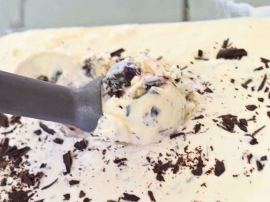 A metal scoop lifts a portion of creamy no churn cherry chip ice cream mixed with chocolate flakes from a dish, with more chocolate shavings on top.