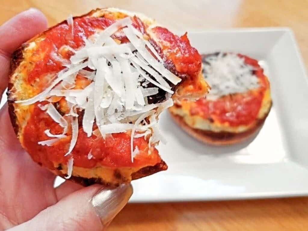 A hand holding a mini pizza with a bite taken out, topped with tomato sauce and shredded cheese&mdash;just like in a classic mini lasagna pizzas recipe. Another mini pizza sits on a white plate in the background.