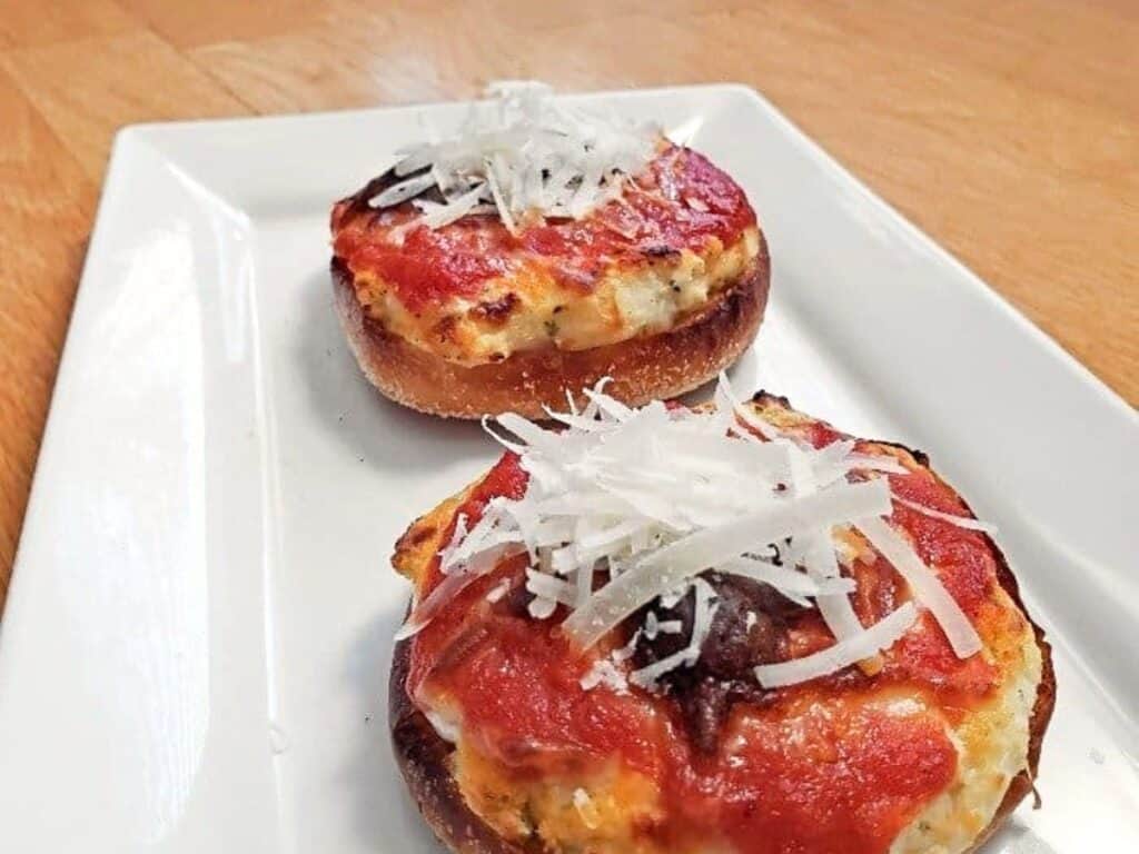 Two mini pizza bagels topped with tomato sauce, melted cheese, and shredded parmesan are served on a rectangular white plate on a wooden surface&mdash;perfect for anyone looking to try a fun twist inspired by the mini lasagna pizzas recipe.