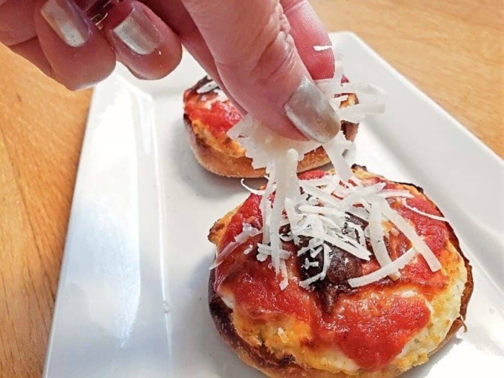 A hand sprinkles shredded cheese onto a mini pizza with tomato sauce, served on a white rectangular plate, perfectly capturing the delicious process of this mini lasagna pizzas recipe.