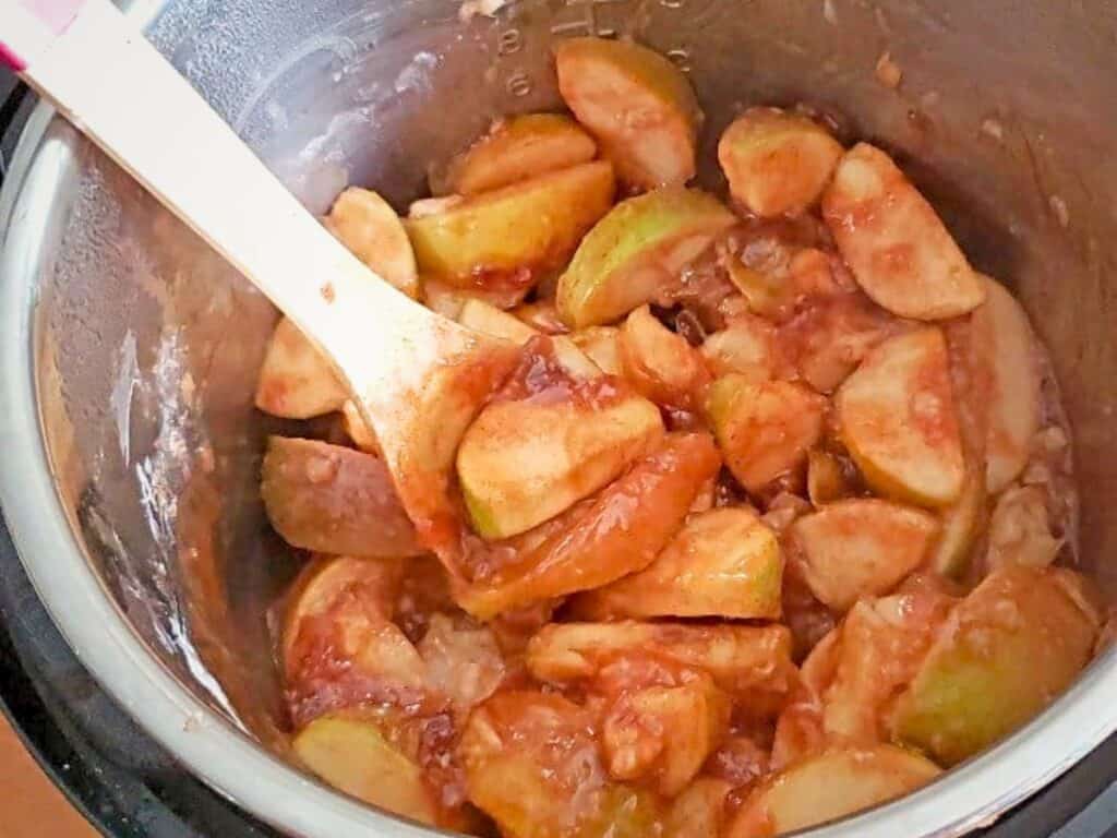 Sliced apples being stirred with a wooden spoon inside a metal pot, likely making homemade Instant Pot cinnamon apples or stewed apples.