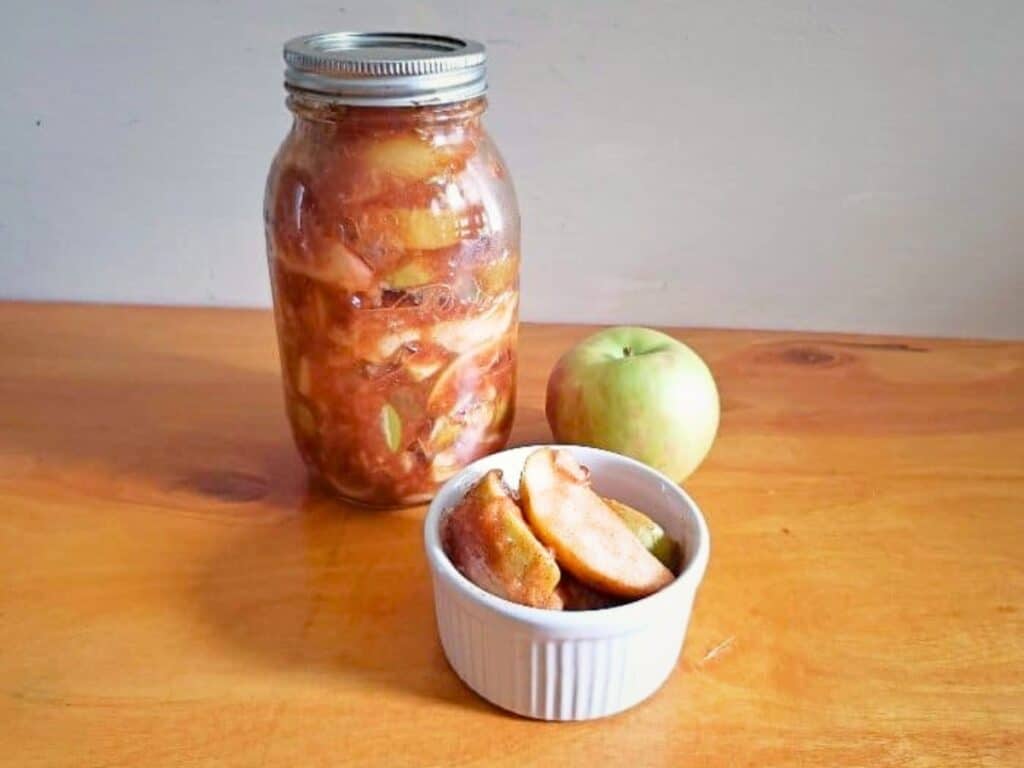 A jar of preserved apple slices, a whole green apple, and a small white bowl with more apple slices&mdash;reminiscent of homemade Instant Pot cinnamon apples&mdash;are arranged on a wooden surface.