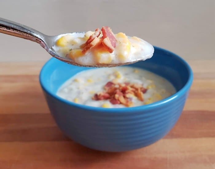 Spoon holding potato and corn chowder with bacon with the full bowl of soup behind it.