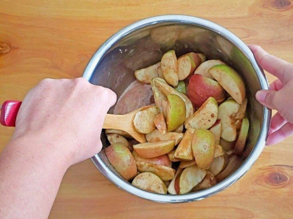 A person stirs sliced apples coated with cinnamon in a metal pot using a wooden spoon on a wooden surface, preparing delicious Instant Pot cinnamon apples.