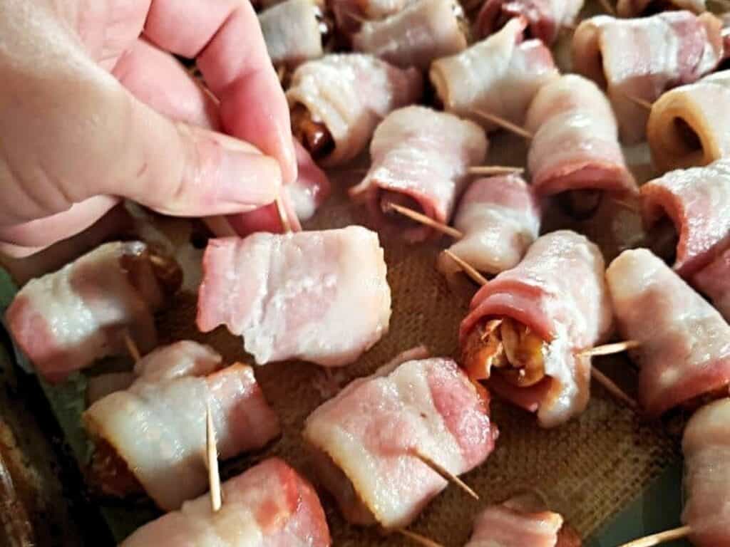 A hand holds a bacon-wrapped appetizer among several others on a tray, each secured with a toothpick&mdash;delicious bacon wrapped dates ready to be enjoyed.