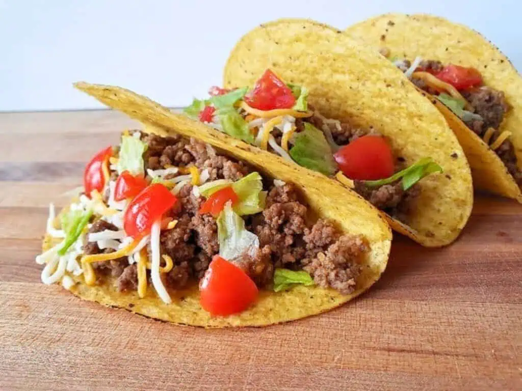 Three hard-shell honey habanero tacos filled with seasoned ground beef, shredded cheese, lettuce, and diced tomatoes on a wooden surface.
