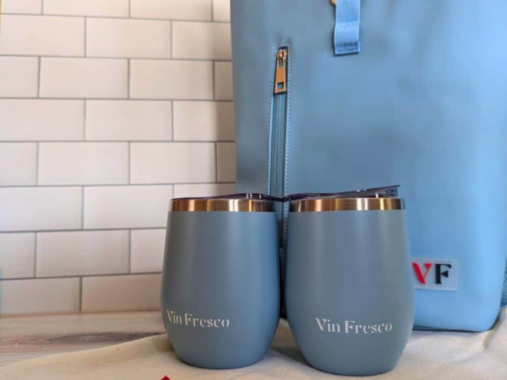 Image shows a light blue wine backpack on a wooden table with two matching tumblers sitting in front of it.