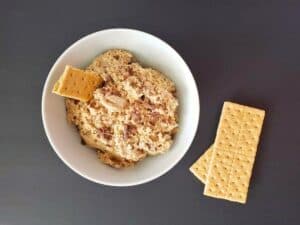 A white bowl filled with creamy edible s’mores cookie dough dip, topped with a piece of graham cracker, sits next to two whole graham crackers on a dark surface.