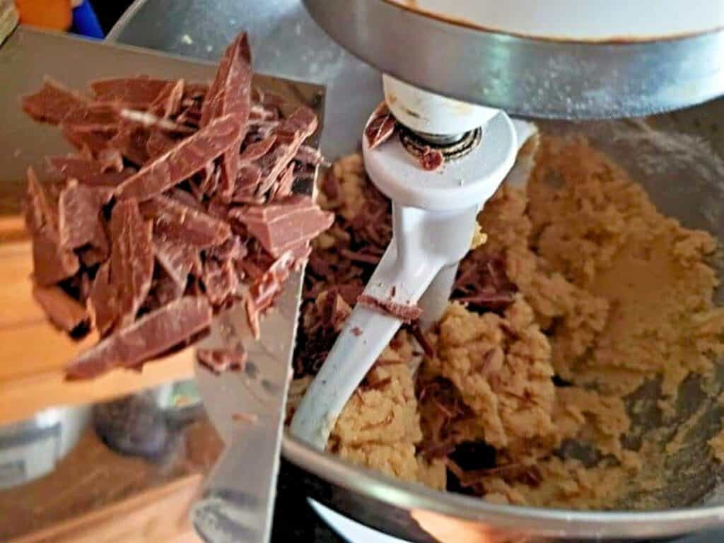 Chocolate shavings are being added to edible s’mores cookie dough in a stand mixer with a white paddle attachment.