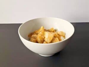 A white bowl filled with white rice is topped with pieces of orange chicken in a light-colored sauce, placed on a dark surface with a plain background.