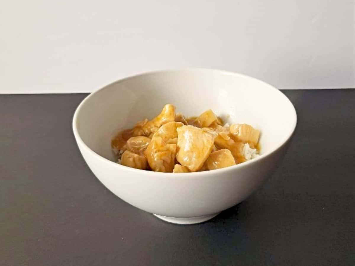 A white bowl filled with white rice is topped with pieces of orange chicken in a light-colored sauce, placed on a dark surface with a plain background.