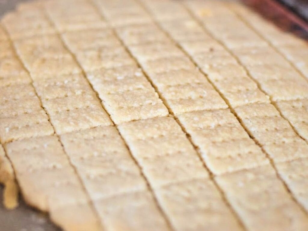 Close-up of a large sheet of baked, golden-brown crackers scored into rectangular pieces with a fork-dotted pattern on top, reminiscent of the precise lines often found on classic peppermint shortbread cookies.