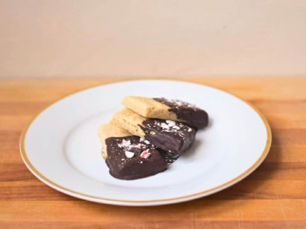 Four peppermint shortbread cookies dipped in chocolate and sprinkled with crushed peppermint are arranged on a white plate with a gold rim, set on a wooden surface.