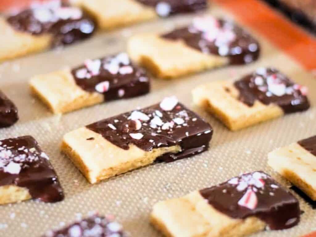Peppermint shortbread cookies in a rectangular shape are partially dipped in chocolate and sprinkled with crushed candy pieces, arranged neatly on a baking mat.