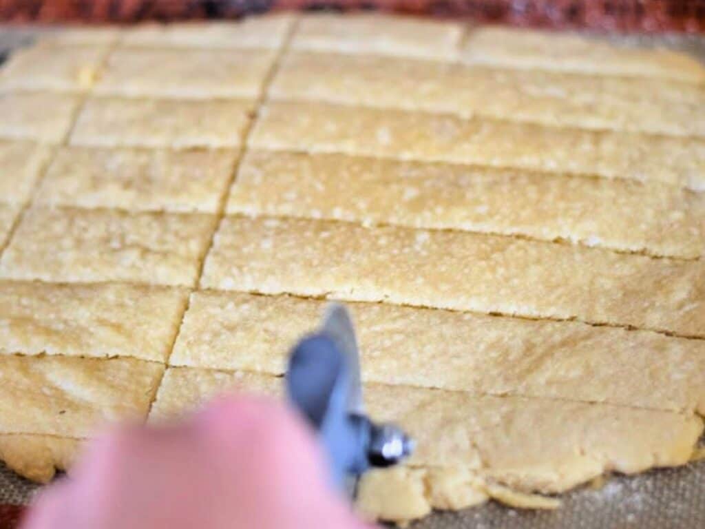 A hand uses a pizza cutter to slice a large sheet of dough into rectangular pieces on a flat surface, preparing the base for peppermint shortbread cookies.