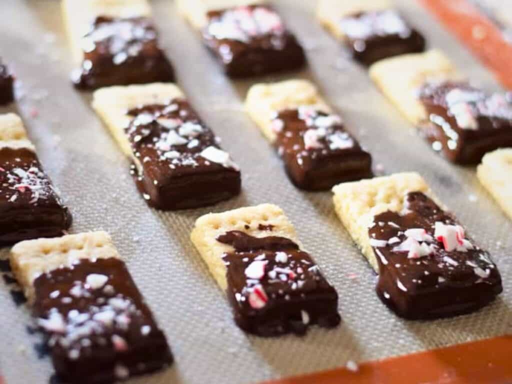 Rows of rectangular peppermint shortbread cookies, partly dipped in chocolate and sprinkled with crushed peppermint, arranged on a baking mat.