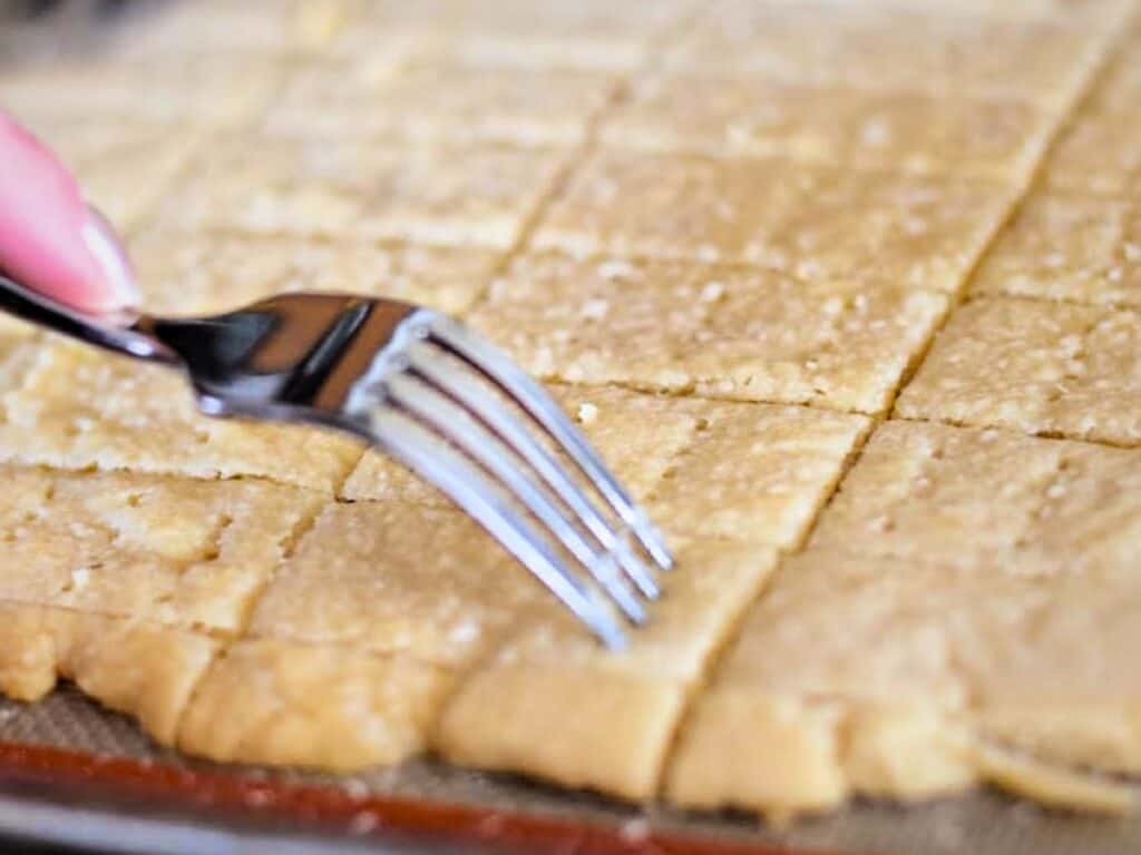 A hand uses a fork to prick holes in a batch of plain peppermint shortbread cookie dough that has been cut into squares on a baking tray.