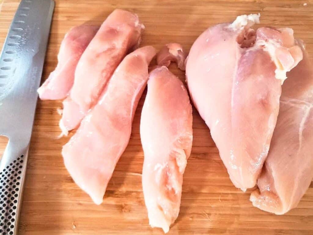 Raw chicken breasts, some whole and some sliced into strips for chicken satay with peanut sauce, are placed on a wooden cutting board next to a metal kitchen knife.