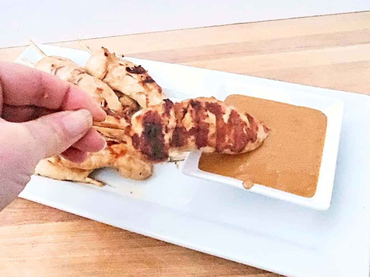 A hand dips a grilled chicken satay with peanut sauce into a bowl of brown sauce on a white rectangular plate, with more skewers in the background.