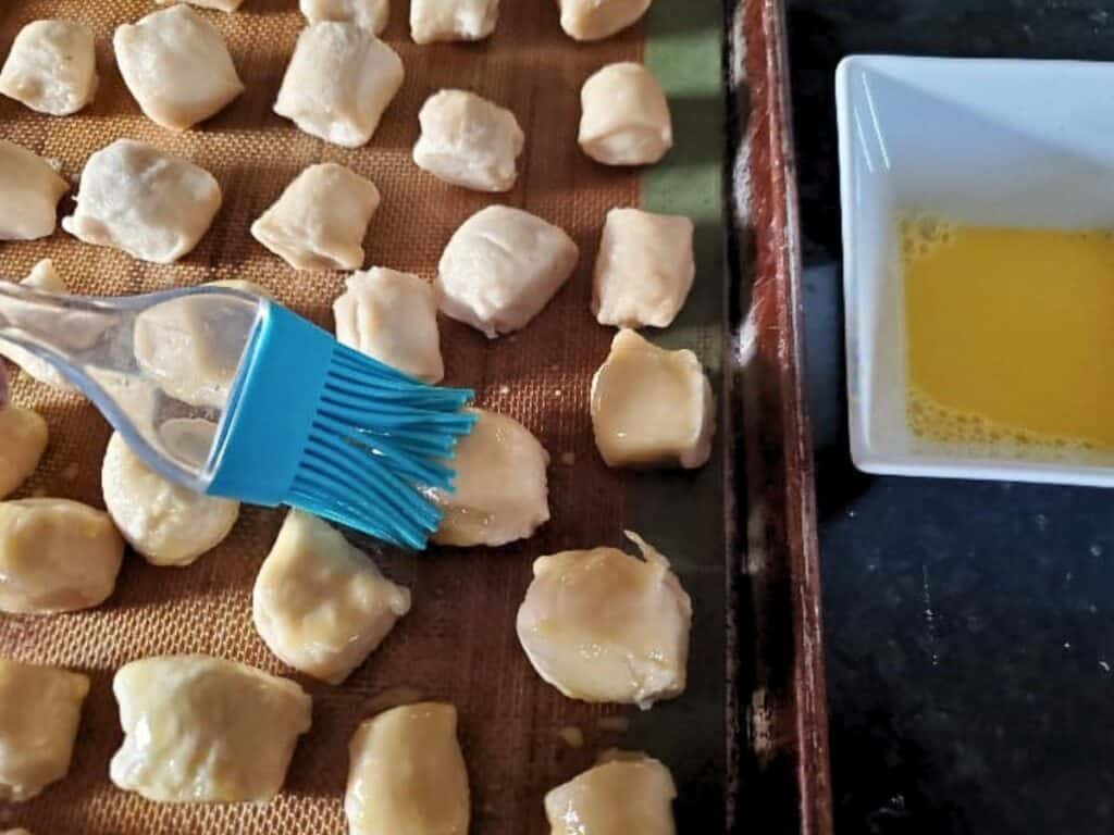 A silicone brush applies egg wash to pretzel bites on a baking sheet; a bowl of egg wash sits nearby.
