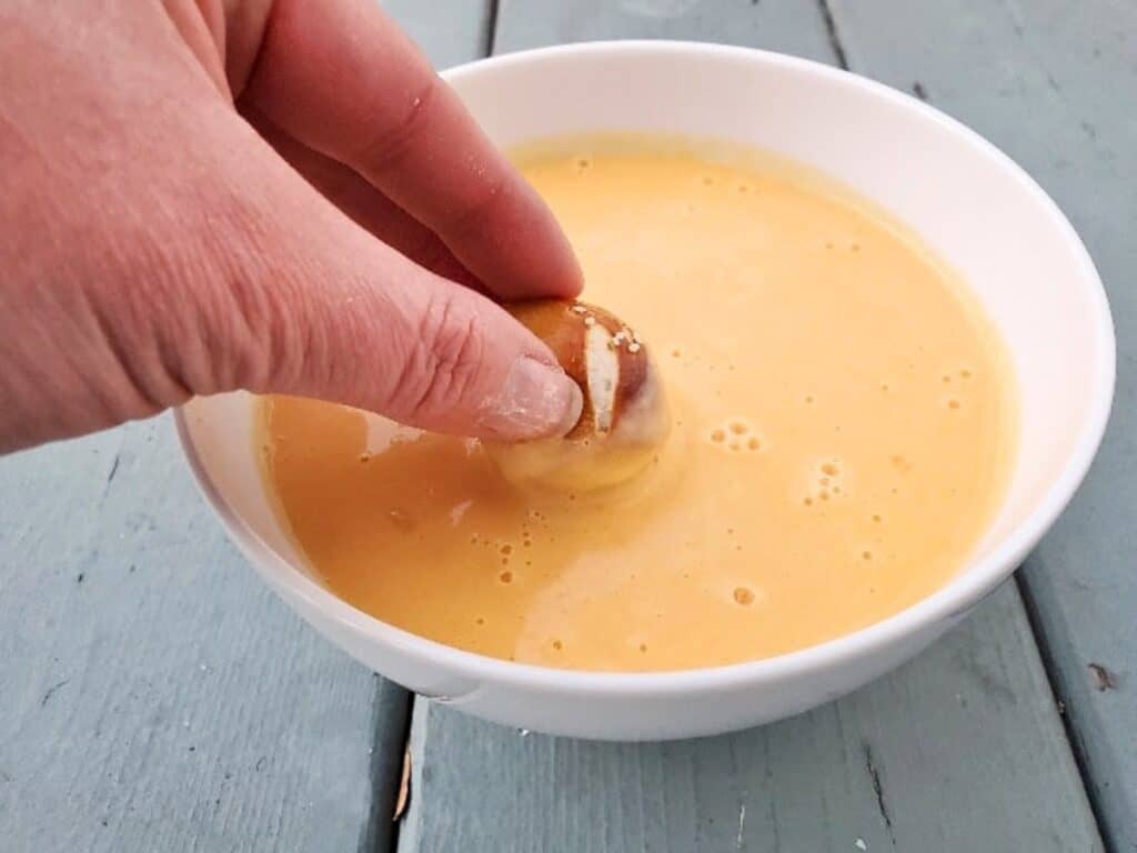 A hand dips one of the soft pretzel bites into a bowl of yellow cheese sauce on a light blue wooden surface.
