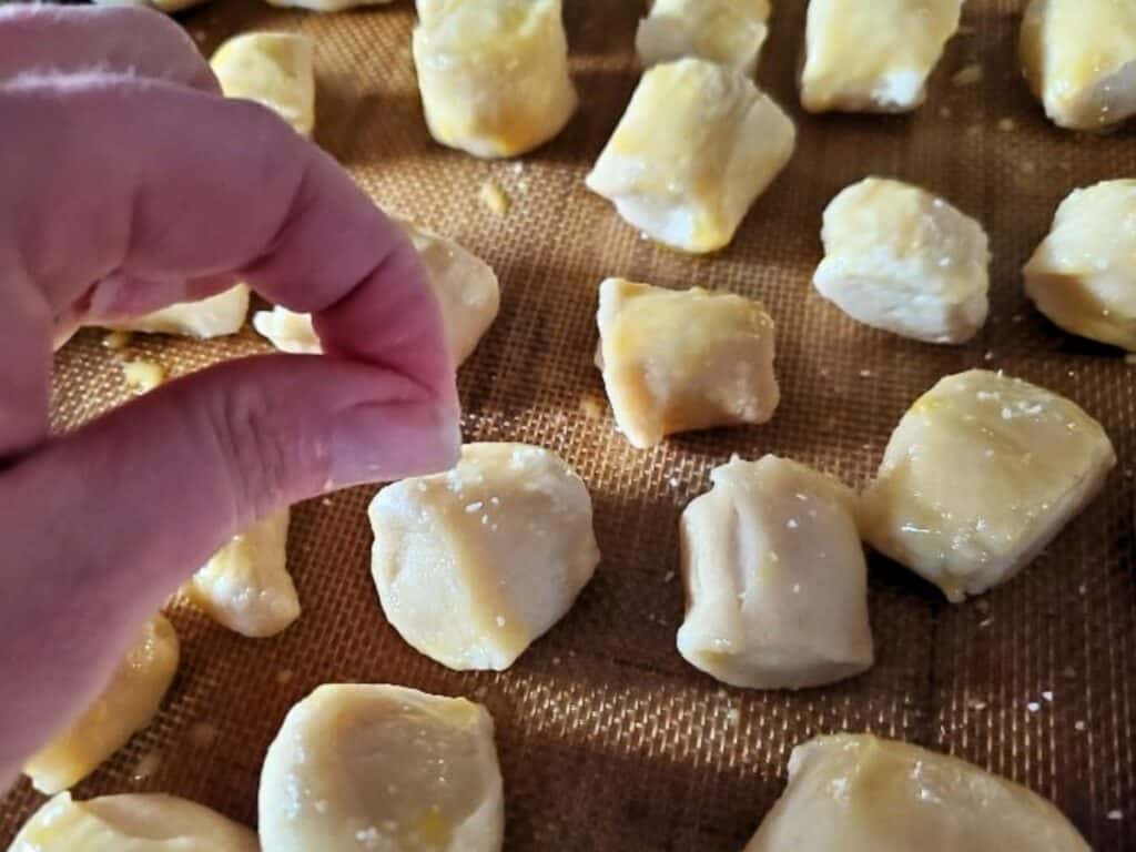A hand sprinkles salt onto unbaked pretzel bites arranged on a baking sheet.