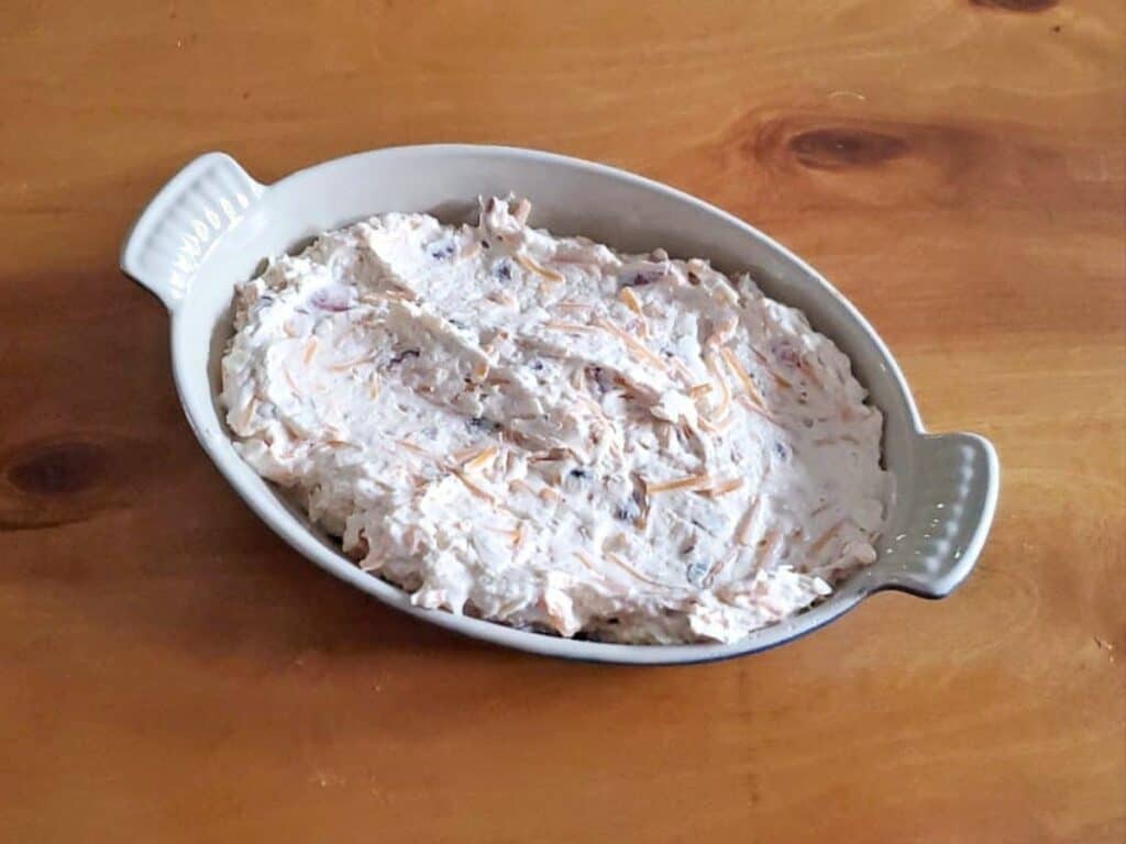 An oval dish filled with a creamy hot bacon cheddar dip, featuring visible shredded cheese and small chopped ingredients, sits on a wooden surface.