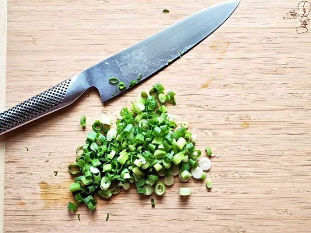 A stainless steel knife lies next to a small pile of sliced green onions on a wooden cutting board, ready to be sprinkled over a savory hot bacon cheddar dip.
