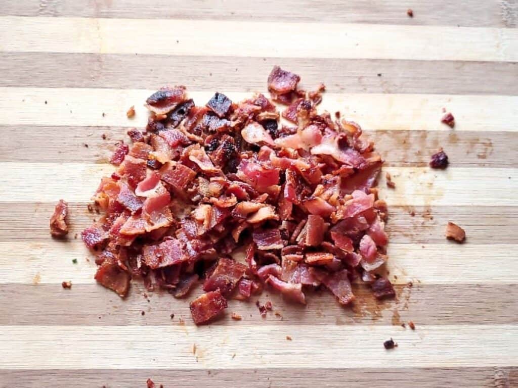 Chopped cooked bacon pieces, perfect for topping a hot bacon cheddar dip, are spread on a striped wooden cutting board.
